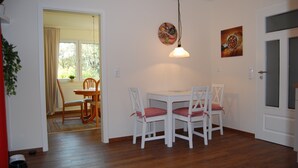 Un coin repas avec une table et des chaises blanches, un luminaire suspendu et une porte sur la droite.