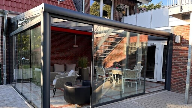 Une terrasse moderne, entièrement vitrée, aménagée avec du mobilier d’extérieur, un mur en briques et un escalier menant à un niveau supérieur.