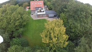 Vue aérienne d’une maison entourée d’une forêt dense, avec une grande pelouse et une construction à deux étages surmontée d’un toit rouge.