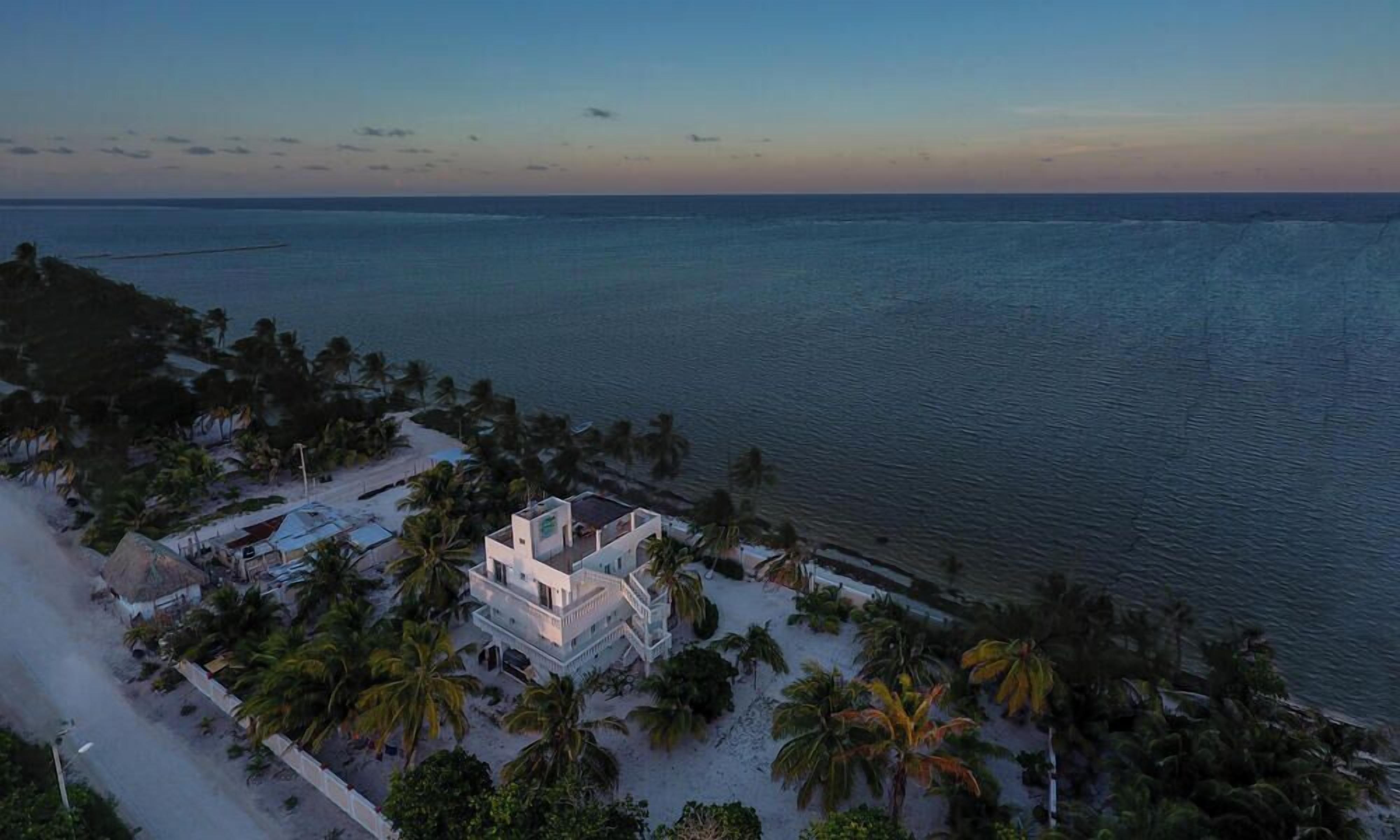 An aerial view of a coastal property with a large building surrounded by palm trees and a beach.