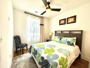 A bedroom with a bed, a chair, a ceiling fan, a window with curtains, and two framed pictures on the wall.