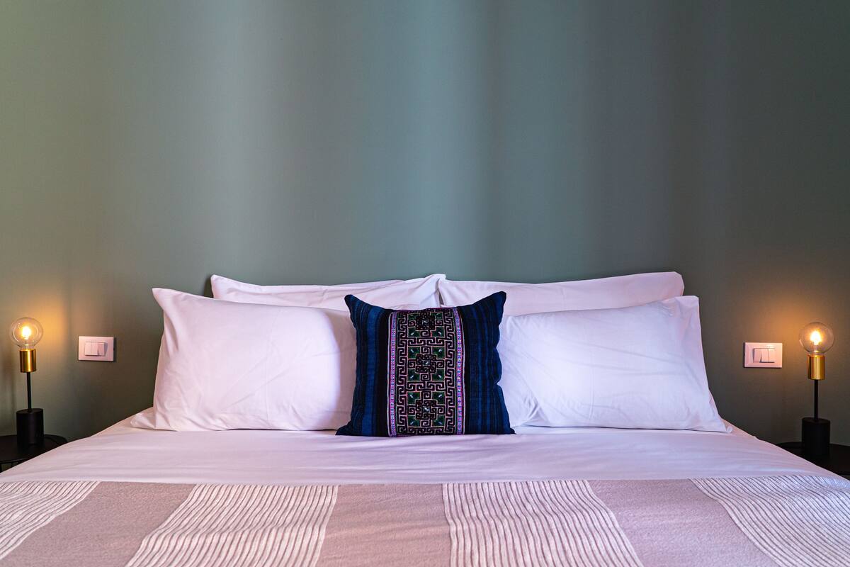 Un lit bien fait, avec du linge de lit blanc et un coussin décoratif, flanqué de deux tables de chevet sur lesquelles sont posées des lampes.