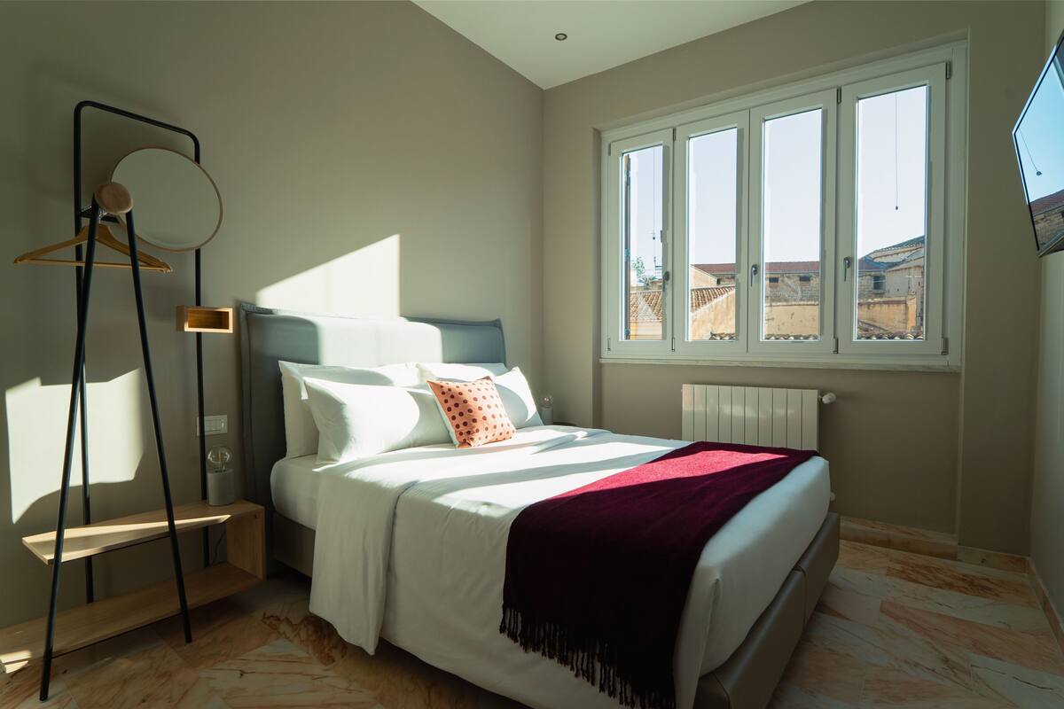 Une chambre à coucher comprenant un lit, une table de chevet, une lampe sur pied, une fenêtre et un téléviseur fixé au mur.