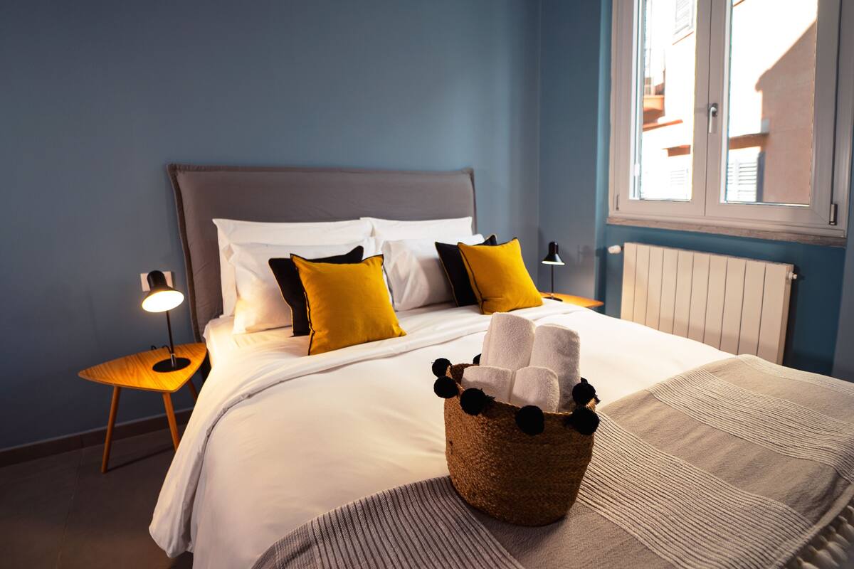 Un lit bien fait, avec du linge de lit blanc et plusieurs oreillers, une lampe de chevet et un panier en osier contenant des serviettes.