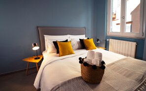 A neatly made bed with white linens and multiple pillows, a bedside lamp, and a woven basket with towels. - Zefiro Suites (Palermo)