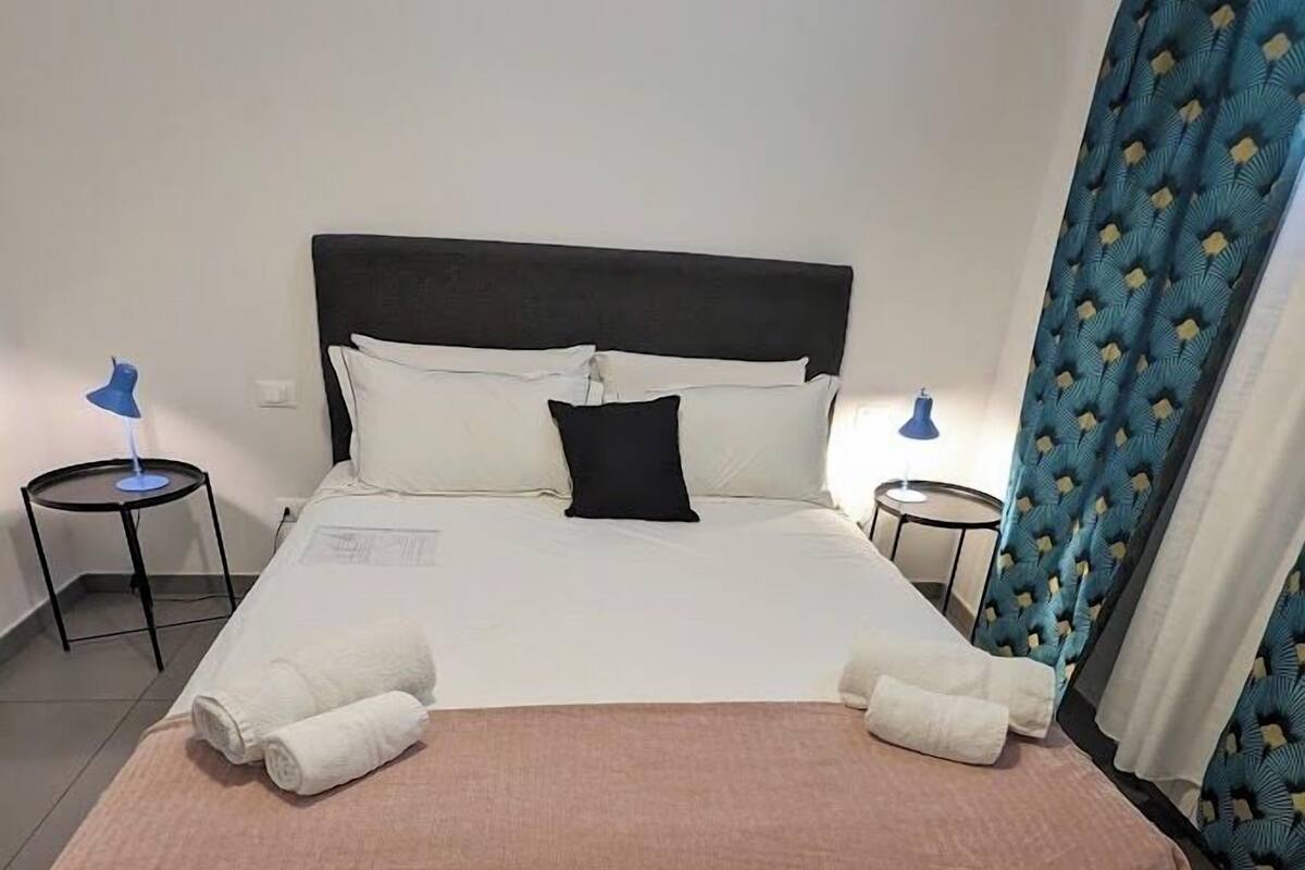 Un lit bien fait, avec du linge de lit blanc et rose, deux tables de chevet avec des lampes et un rideau bleu.