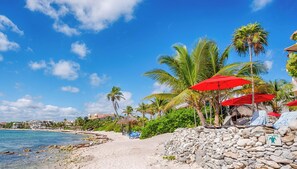 Ein Strand mit Palmen, einer Steinmauer und roten Sonnenschirmen.