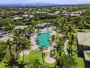 An aerial view of a resort with a central swimming pool, surrounded by palm trees and houses.