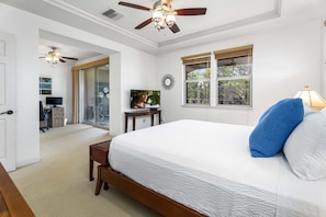 A bedroom with a large bed, a ceiling fan, a window with a view, and a door leading to another room.