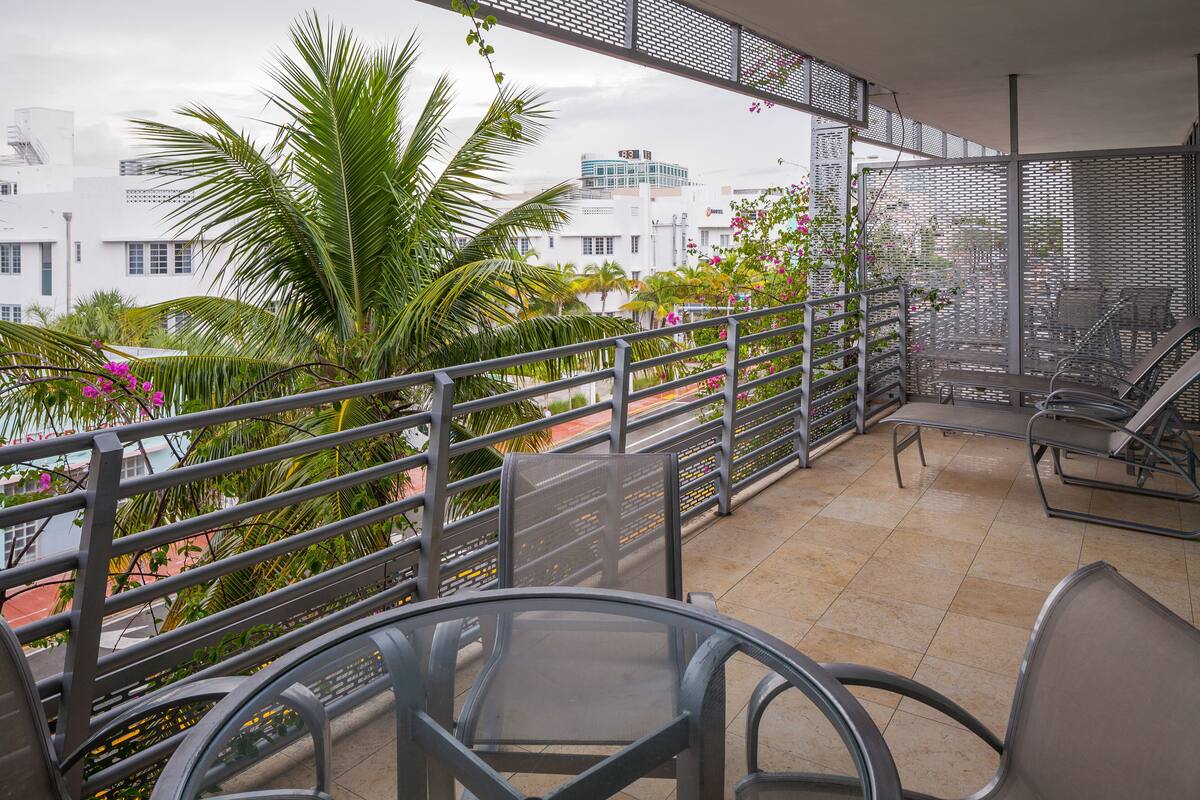 Un balcón con barandilla de metal, una mesa de vidrio y sillas. Hay plantas y flores alrededor, y un paisaje urbano al fondo.