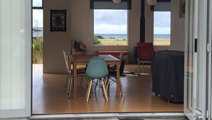 An open-plan living area with a dining table, chairs, and a sofa. There are large windows offering a view of the outdoors. - Wairarapa Bach Flat Point (Flat Point)
