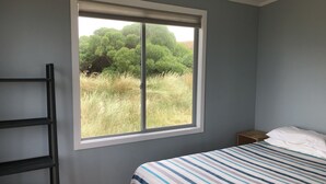 A bedroom with a bed, a window with a view of greenery, and a black shelf unit. - Wairarapa Bach Flat Point (Flat Point)