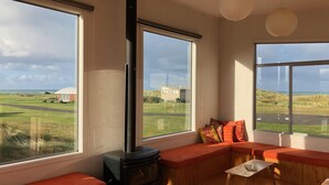 A cozy room with a wood-burning stove, large windows offering views of the countryside, and a seating area with cushions. - Wairarapa Bach Flat Point (Flat Point)