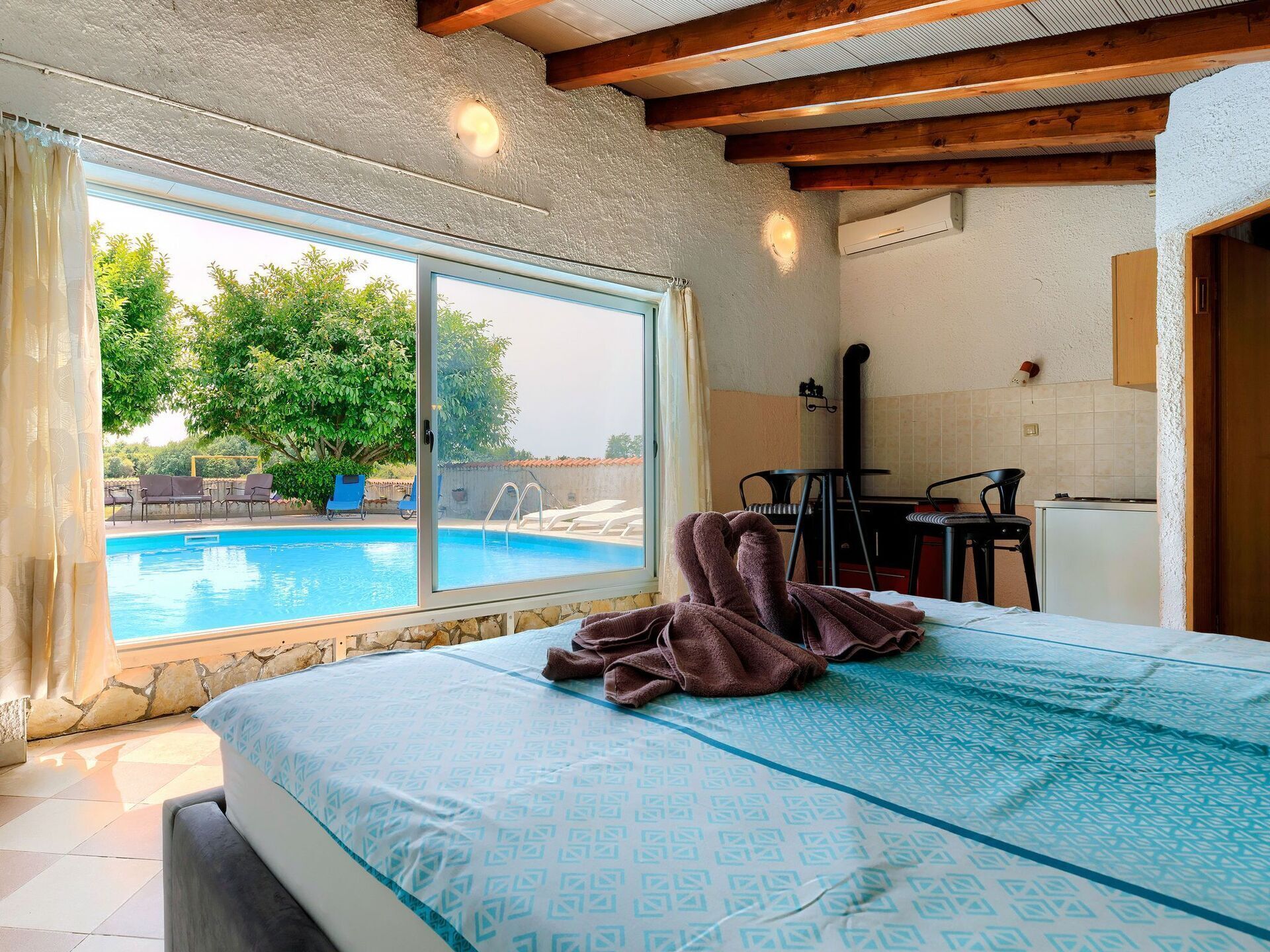 A bedroom with a large window overlooking a swimming pool, a bed with a patterned bedspread, and a kitchenette area.