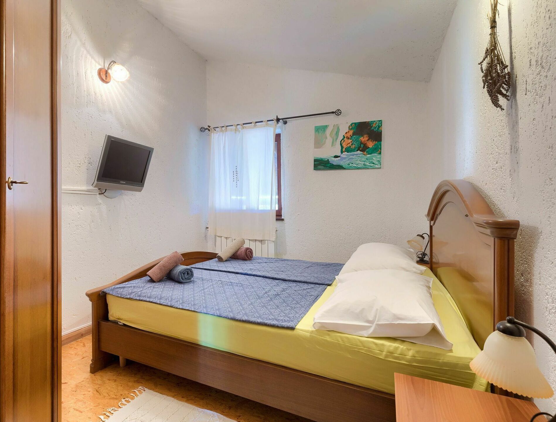 A bedroom with a bed, a television, a window with curtains, and a painting on the wall.