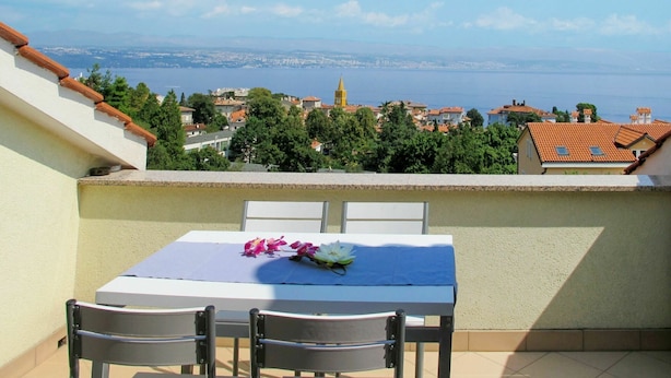 A balcony with a table and chairs, overlooking a coastal town with a clear view of the sea and distant cityscape.