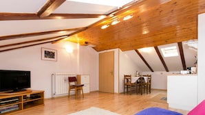 A cozy attic room with a wooden ceiling, a TV, a dining table, and a sofa.