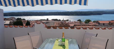 A table with chairs under a striped awning, overlooking a lake with buildings in the background.