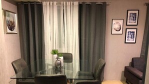 A modern living room with a glass table and two chairs, a sofa, and a large window with curtains.