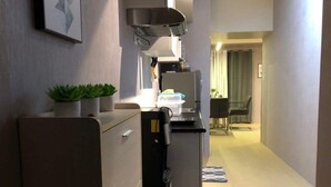 A compact kitchen with a refrigerator, microwave, and a small countertop. A potted plant is on the counter, and a dining area is visible in the background.