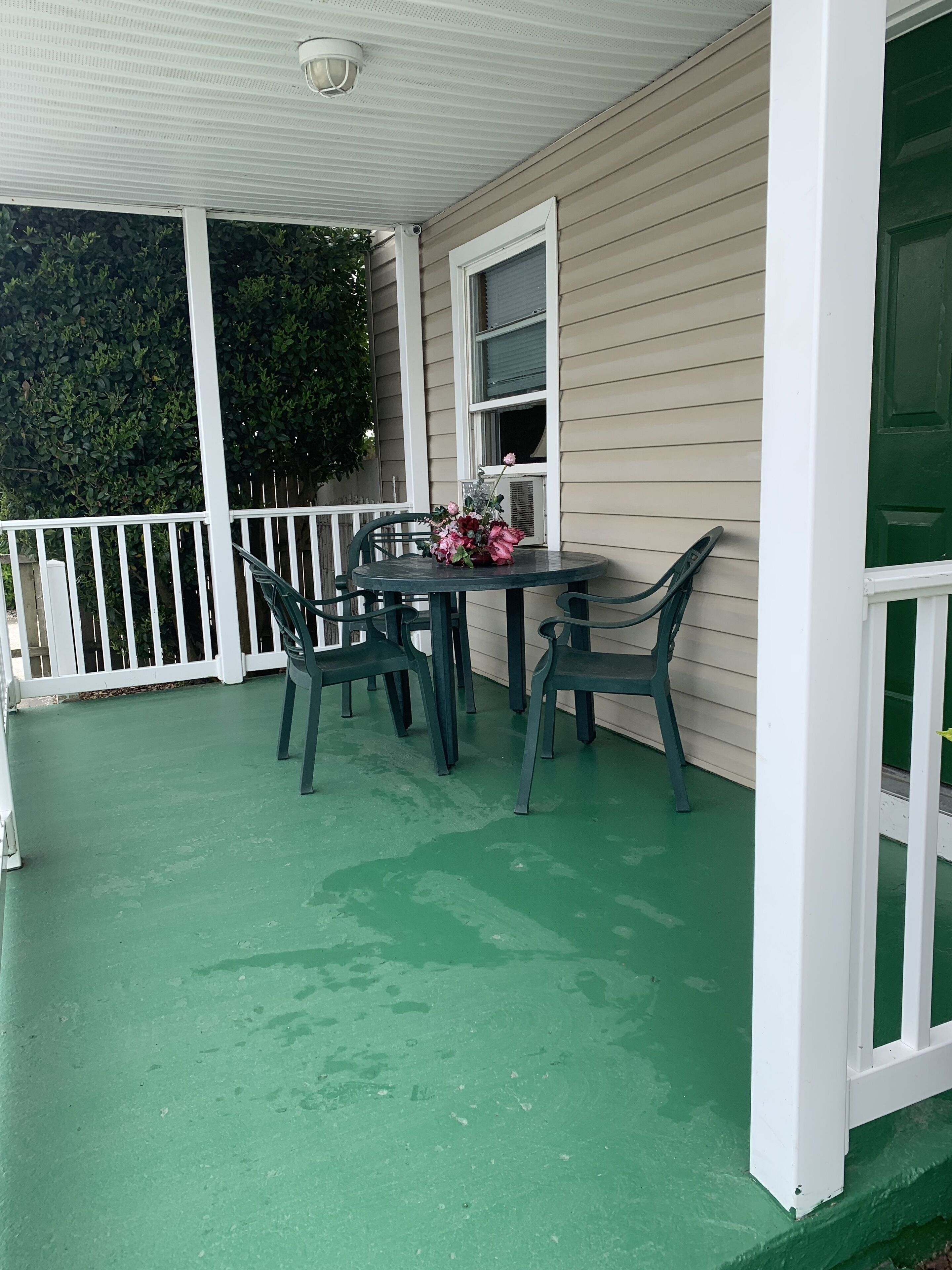 Een overdekte veranda met een groene vloer, een tafel met stoelen en een raam met bloemen.