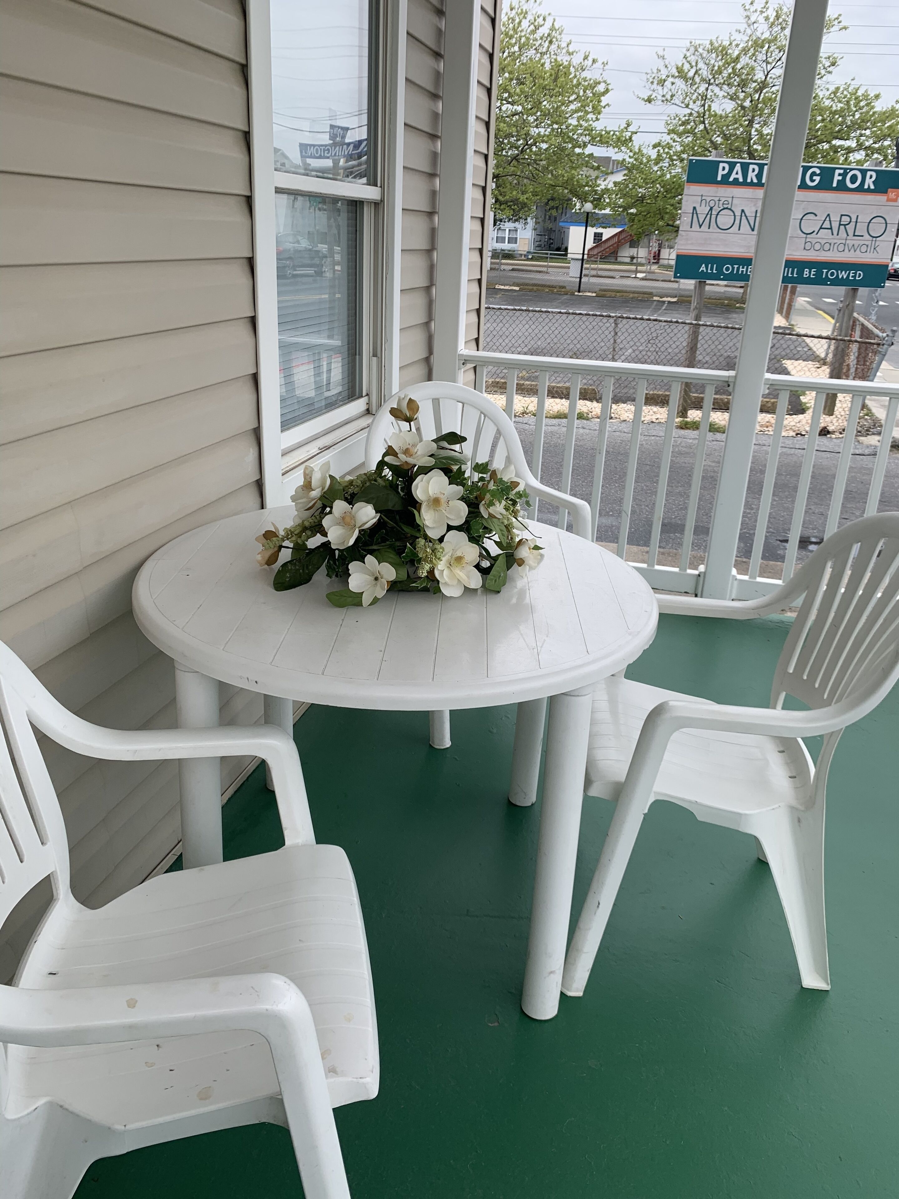 Een klein buitenzitgedeelte met een ronde tafel en witte plastic stoelen, een bloemstuk op de tafel, en een bord dat parkeergelegenheid aangeeft voor hotel Mon Carlo boardwalk.