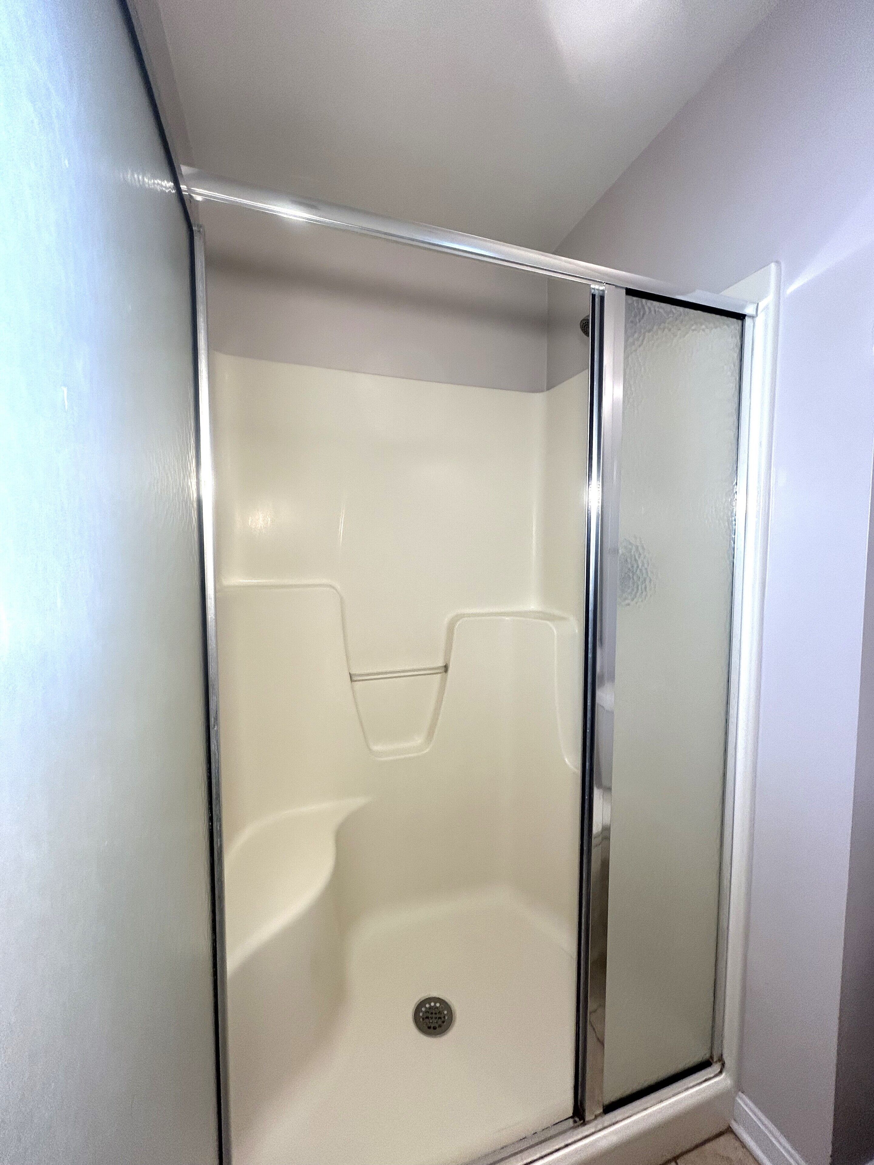 A glass-enclosed shower stall with a white interior and a drain in the center.