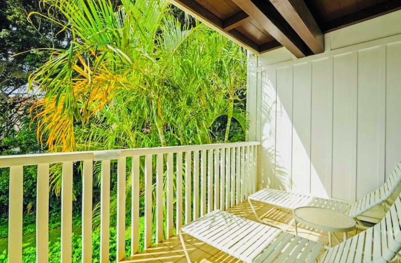 A covered outdoor seating area with white metal chairs and a small round table, surrounded by lush green foliage.
