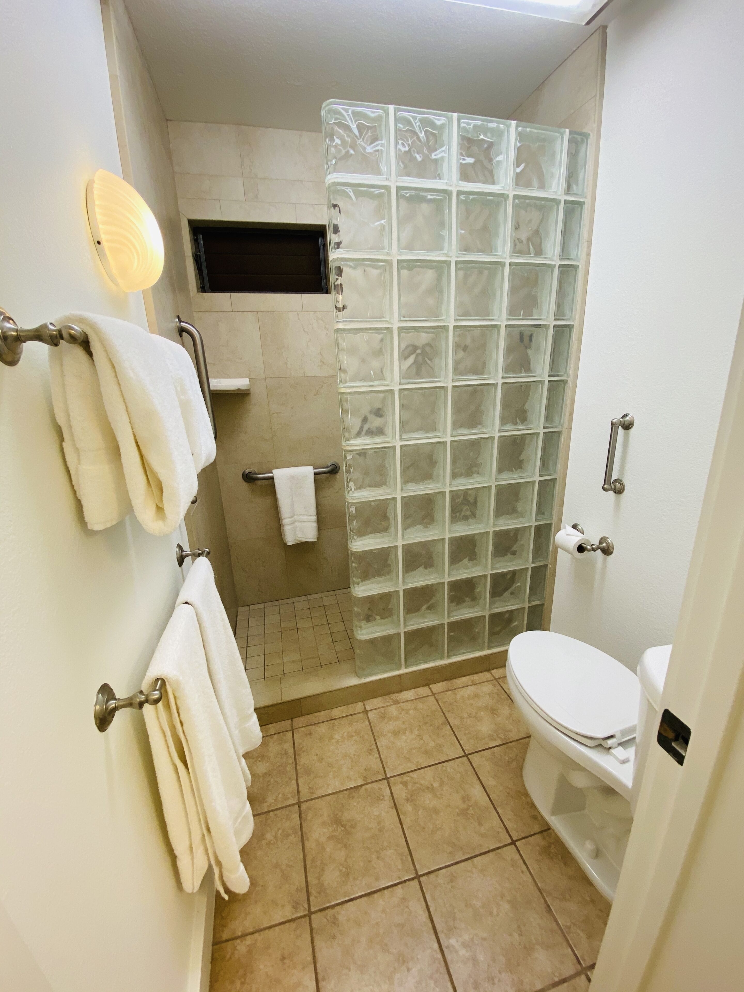 Un baño con inodoro, cabina de ducha de cristal, toallero con varias toallas, barra de apoyo y un aplique en la pared.