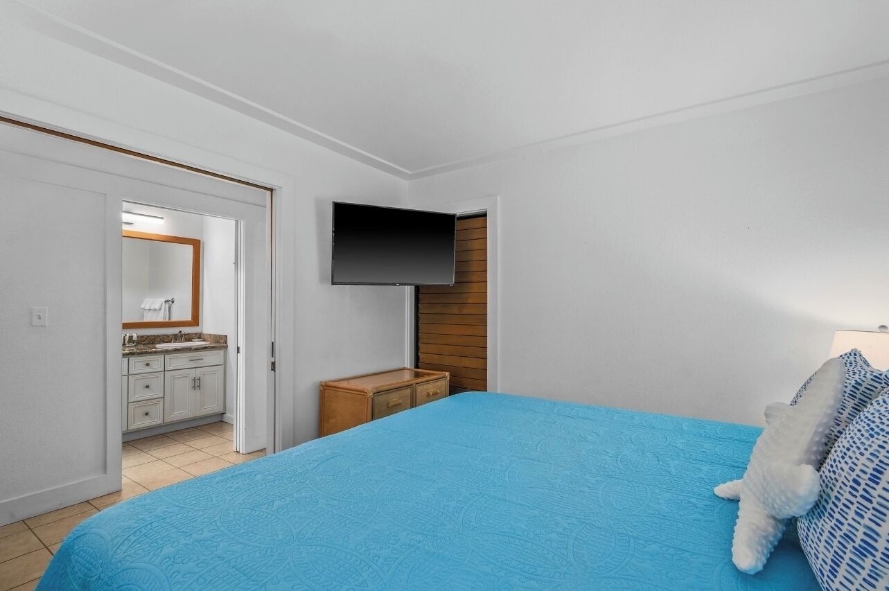 Une chambre à coucher avec un lit, un téléviseur fixé au mur, une table de chevet en bois et une salle de bain visible à travers une porte ouverte.