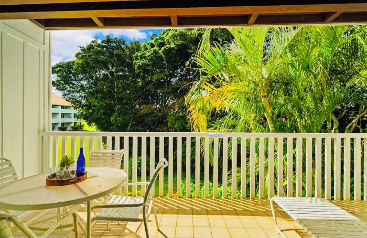 A covered outdoor area with a table and chairs, surrounded by a white fence and lush greenery.