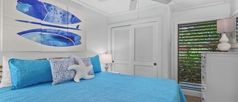 A bedroom with a large bed, a ceiling fan, a window with blinds, and a nightstand with a lamp.