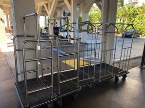 Carrinhos de metal com rodas, provavelmente para bagagem ou equipamentos, sob uma área coberta com pilares de concreto.
