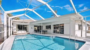 Una piscina amb aigua transparent i blava, envoltada d'una estructura blanca amb portes i finestres de vidre, i un sostre amb llums penjants.