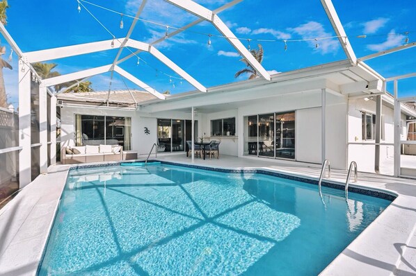 Una piscina con agua azul cristalina, rodeada por una estructura blanca con puertas y ventanas de vidrio, y un techo con luces colgantes.