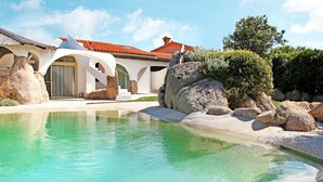 Una villa moderna con piscina, grandes rocas y techo de tejas rojas.