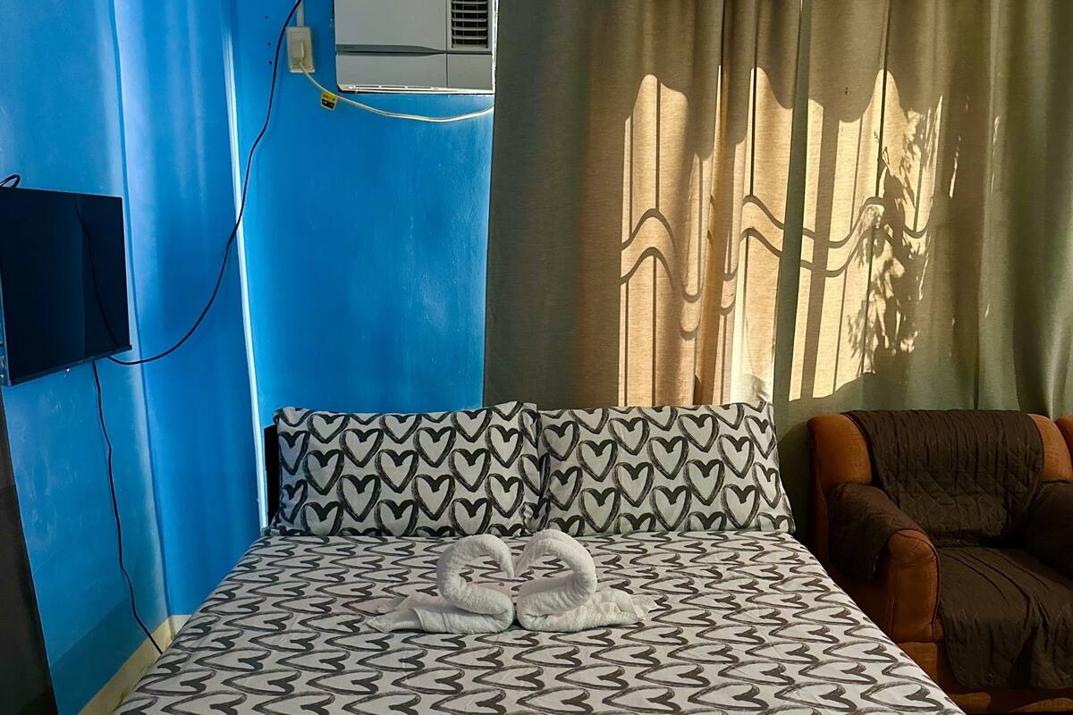 A hotel room with a patterned bedspread, a wall-mounted air conditioner, and a view of the outdoors through curtains.