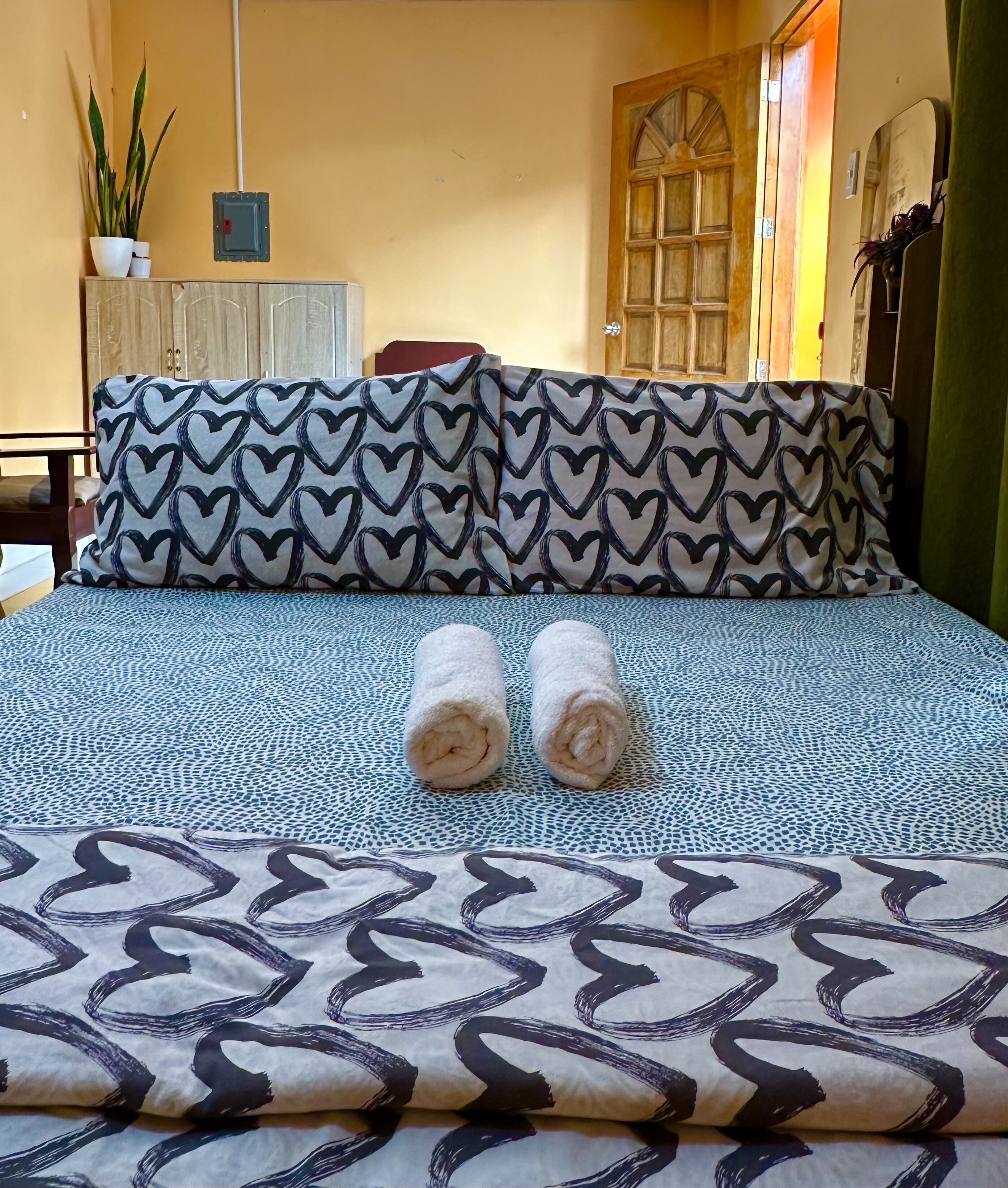 A bed with patterned bedding, two rolled towels, a potted plant, and a wooden door.