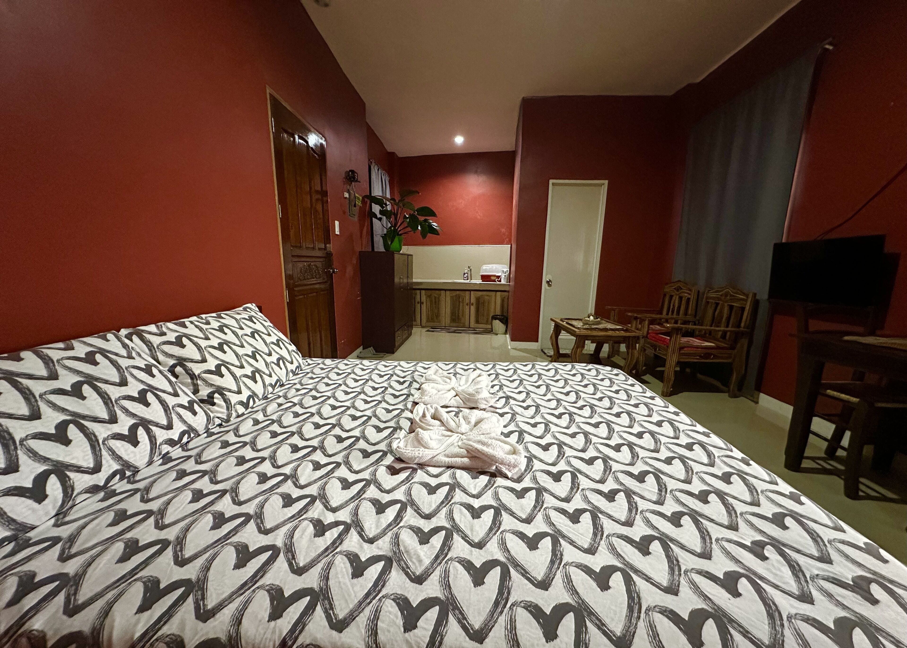 Habitación de hotel con una cama grande, una mesa de comedor, un televisor y una kitchenette al fondo.