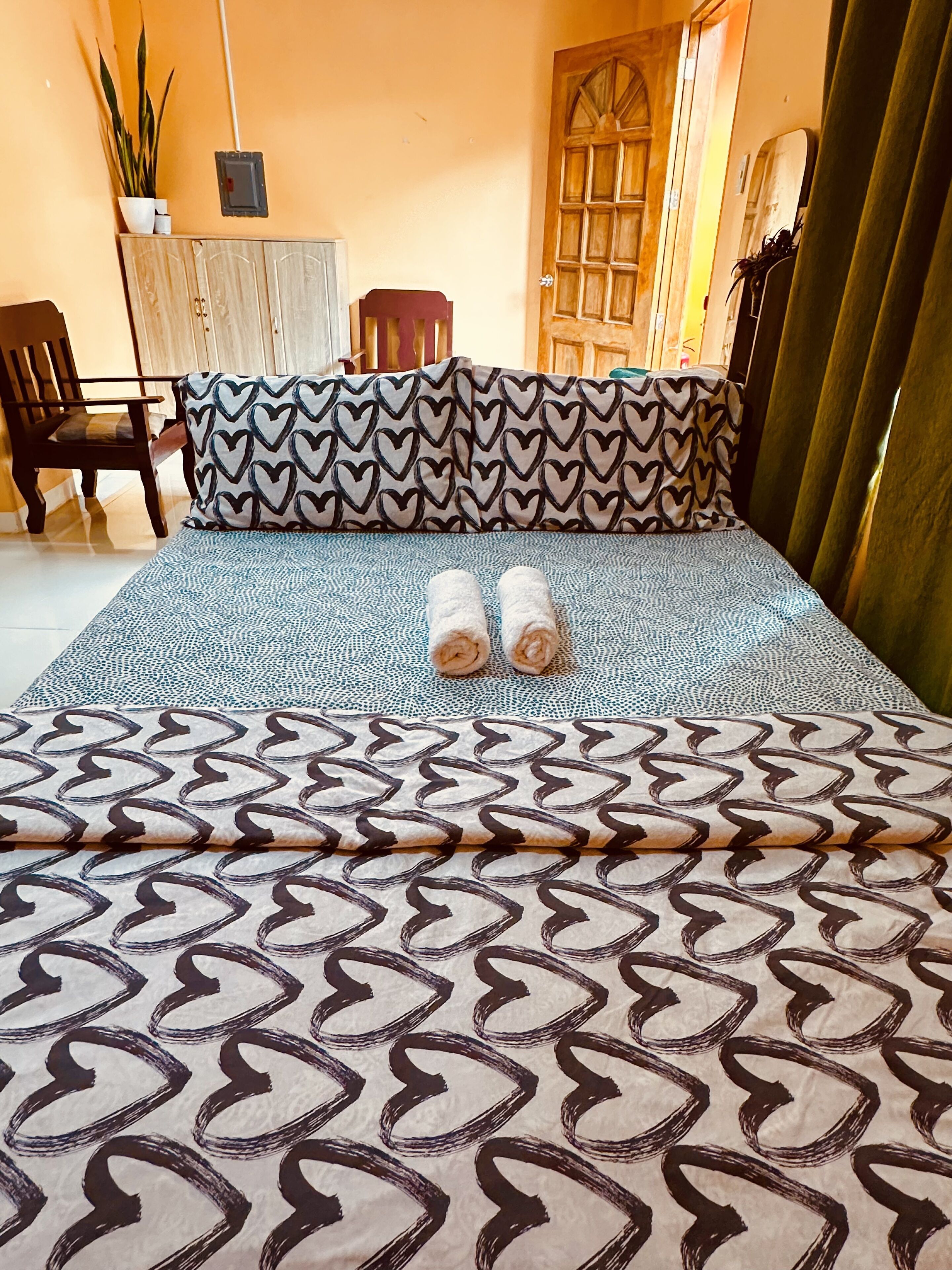 Una cama bien hecha con ropa de cama estampada, una silla de madera, un escritorio con una planta y una puerta con ventanal.