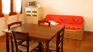 A dining area with a wooden table and chairs, a sofa with a patterned cover, a television on a cabinet, and a window with curtains.