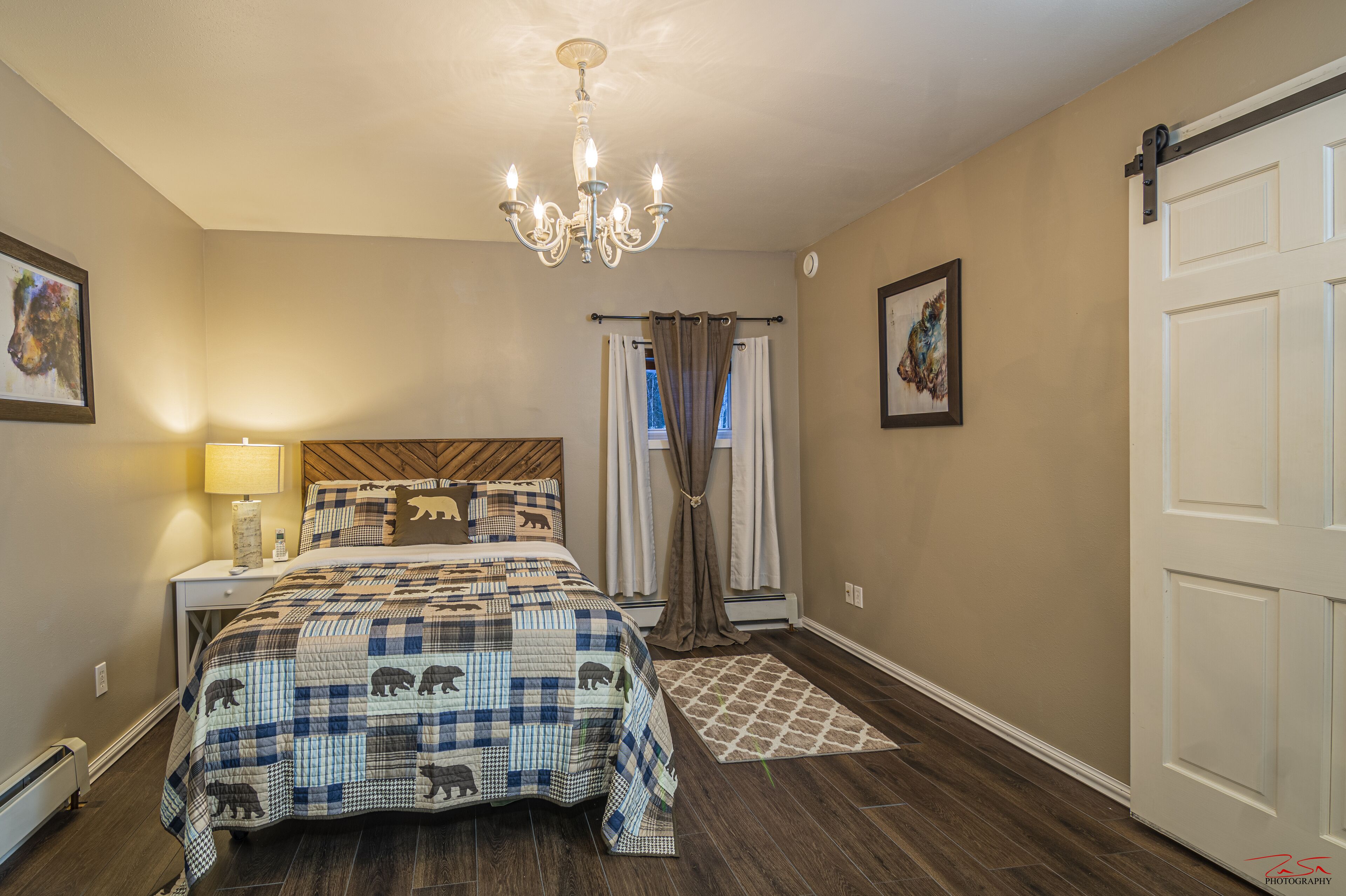 Quarto com cama, mesa de cabeceira, abajur, lustre, janela com cortinas e porta.