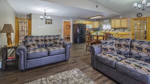 A living room with two sofas, a coffee table, and a rug. There is a kitchen in the background with wooden cabinets and a refrigerator.