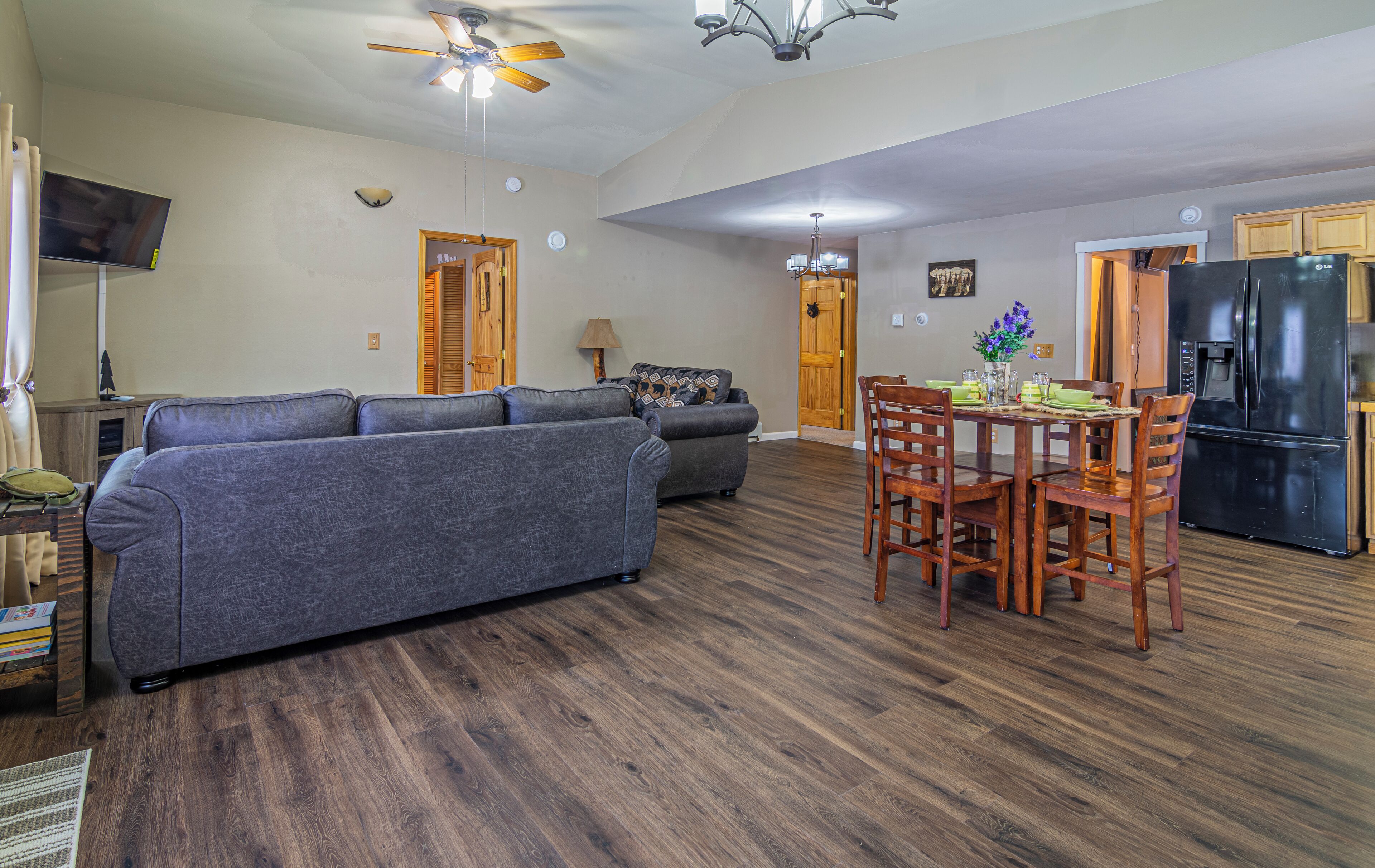 Ein großzügiges Wohnzimmer mit grauem Sofa, ein Essbereich mit Holztischen und einer Tafel, und eine Küche mit einem schwarzen Kühlschrank.