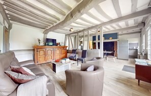 A spacious living area with a high ceiling, wooden beams, a fireplace, and a kitchen in the background.