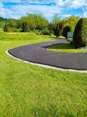 A winding paved path in a green landscape with a vehicle parked on the side. - Homerez - House in Vittefleur, garden. (Vittefleur)
