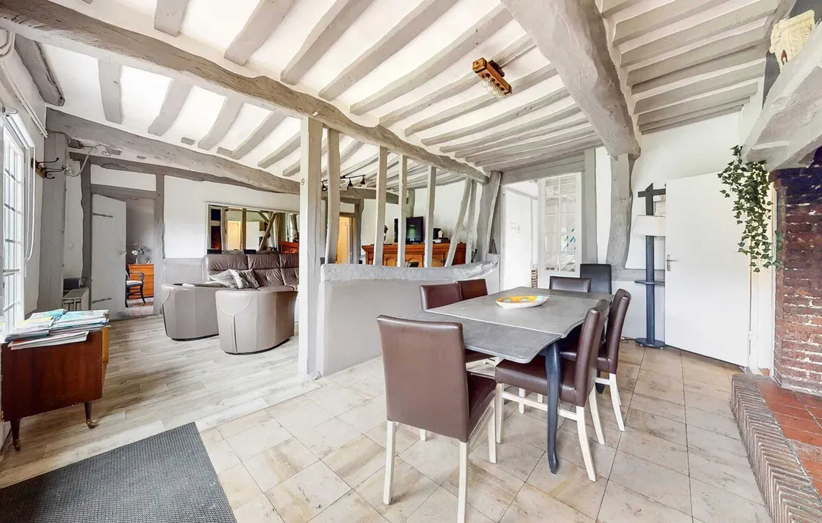 Un vaste espace dédié à la salle à manger, avec une grande table et des chaises, une cuisine à l’arrière-plan et un plafond haut avec des poutres apparentes.