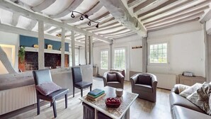 Ein geräumiges Wohnzimmer mit hohen Decken, freiliegenden Balken und großen Fenstern. Es gibt mehrere Sitzbereiche mit Ledersofas und -sesseln.