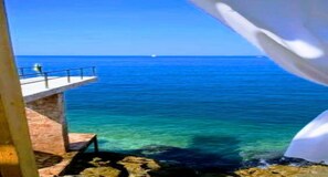 Une vue sur la côte, avec une mer bleue et limpide, une jetée qui s’avance dans l’eau et une côte rocheuse.