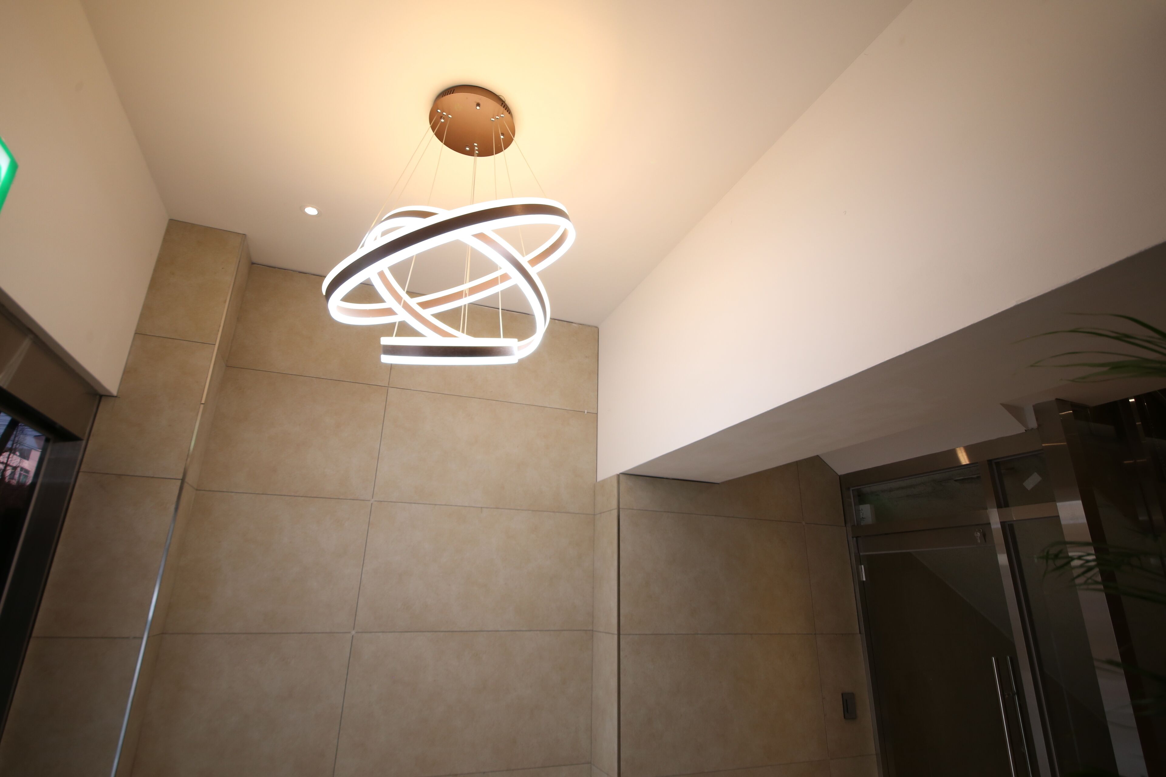 A modern interior with a unique ceiling light fixture, a glass elevator, and a beige tiled wall.